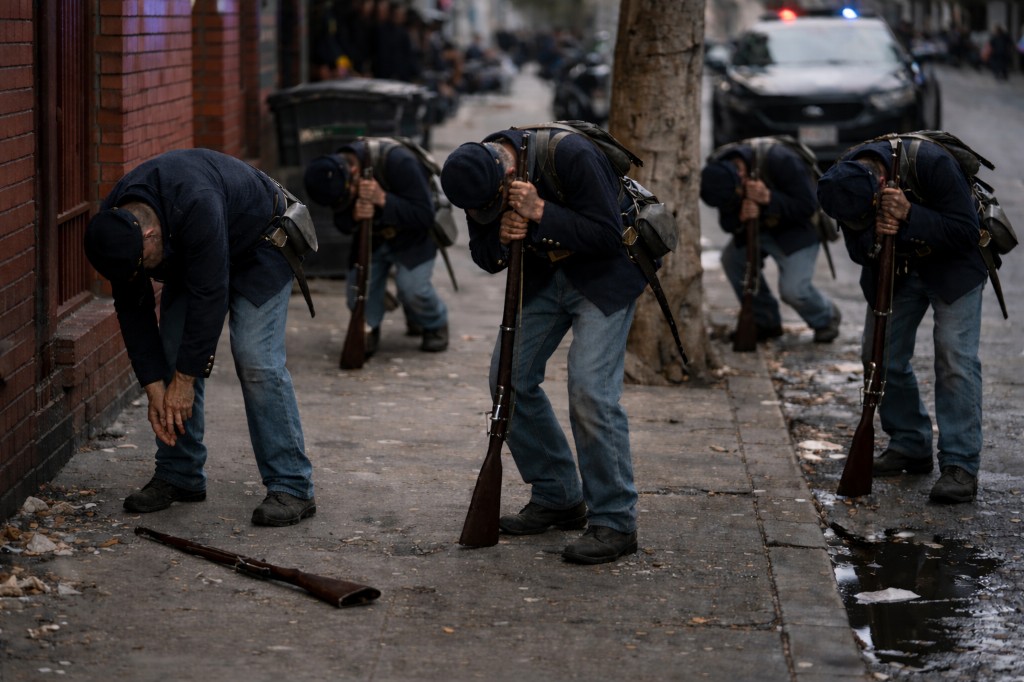 Five people in Union Civil War uniforms on a sidewalk; four lean forward on upright rifles with heads bowed, one bends at the waist with a rifle on the ground; brick wall and street behind them.